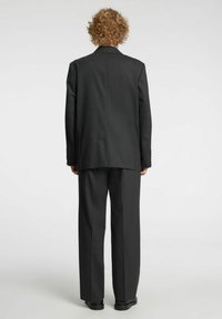 Giacca da completo nera con vestibilità comoda, rever a lancia e due spacchi posteriori, abbinata a pantaloni a gamba larga coordinati e scarpe basse.