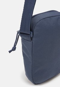 Marinsyndig tygaxelväska med en texturerad yta, rektangulär form och justerbar rem. Stängs med dragkedja och har en dragsnöre.