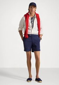 Ναυτικές βερμούδες με λευκό κουμπί, λευκό πουκάμισο από λινό, κόκκινο πλεκτό πουλόβερ τοποθετημένο στους ώμους και ναυτικές εσπαντρίγιες. Ριγέ μπλούζα από κάτω.