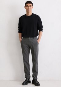 Black sweater, gray textured trousers, black lace-up shoes; model stands with hands in pockets against a light background.