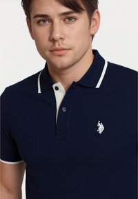 Young man with short dark hair is wearing a navy polo shirt with white collar and a logo. He has a neutral expression and is facing forward.
