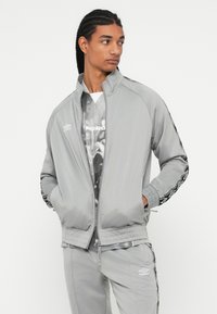 Persona joven con trenzas, vistiendo un chándal gris de Umbro, con las manos en los bolsillos, mirando de lado contra un fondo blanco liso.