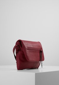 Červená kožená kabelka přes rameno s klopou přehnutou přes přední část, boční kapsou na zip a nastavitelným popruhem. Hladká textura s minimálním šitím.