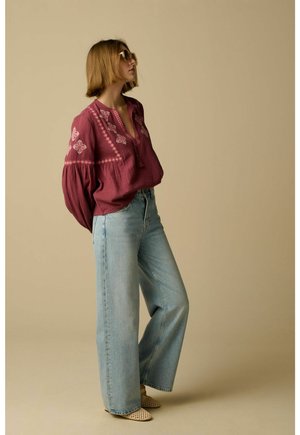 Femme portant des lunettes de soleil rondes, un chemisier brodé bordeaux, un jean large bleu clair et des chaussures tissées beige, debout devant un fond uni.