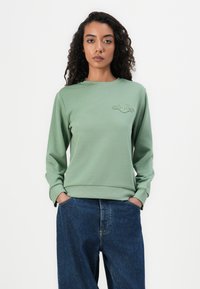 Felpa in cotone verde con scollatura rotonda, polsini a coste e un piccolo logo ricamato sul petto, abbinata a jeans di denim larghi.