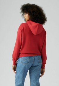 Felpa rossa con cappuccio regolabile e maniche a costine, abbinata a jeans azzurri a vita alta. Texture liscia, design con vestibilità rilassata.