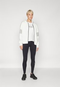 Bijela zip-up majica s crnim prugama na rukavima, bijela majica s grafikom, crne tajice i crne atletske tenisice.