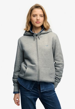 Kvinde iført en lysegrå hættetrøje med lynlås over en blå denims skjorte og blå jeans, stående mod en ensfarvet lys baggrund.