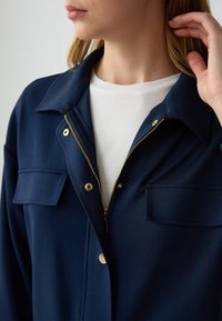 Giacca blu navy con zip anteriore, accessori dorati e due tasche sul petto. Texture morbida, colletto classico, indossata sopra una camicia bianca.