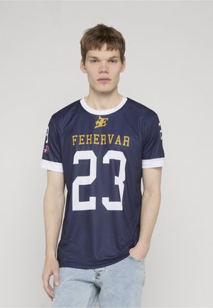 Ung mand iført en mørkeblå sportstrøje med "FEHERVAR" og nummer 23, stående med den ene hånd i lommen på lyseblå jeans.