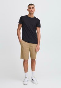 Svart bomull crew-neck t-skjorte, khaki shorts med lommer, hvite sokker, og sorte og hvite joggesko. Modell står mot en ensfarget bakgrunn.