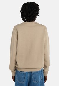 Person with dark dreadlocks wearing a beige long-sleeve sweatshirt and blue denim jeans, viewed from the back against a plain background.