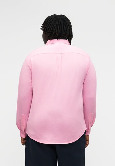 Person med mørk hud og dreadlocks iført en langermet rosa skjorte og mørke bukser, sett bakfra mot en ensfarget bakgrunn.