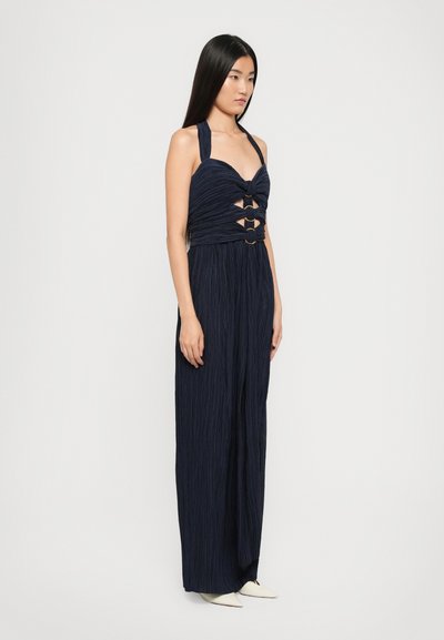 Navy pleated jumpsuit with a halter neck, featuring cutouts secured by round gold hardware, and tapered legs, with a smooth texture.