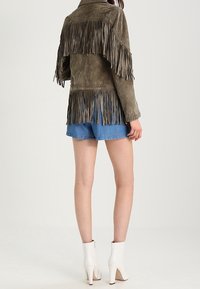 Femme portant une veste en suède frangée marron, un short en denim bleu et des bottines à talons hauts blanches, se tenant devant un fond uni.
