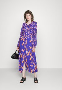 Vestido longo floral roxo com padrões laranja e azul; apresenta mangas longas, decote redondo e uma abertura frontal. Usado com mules pretas e uma bolsa.