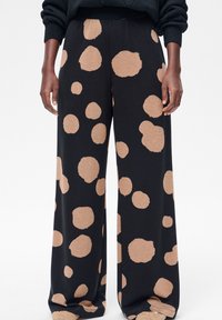 Zwarte wijdvallende broek met grote onregelmatige beige polkadots, gedragen met een zwarte trui en beige pantoffels.