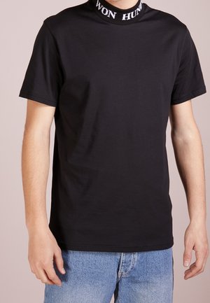 Homme portant un t-shirt noir uni à manches courtes avec « WON HUND » imprimé sur le col roulé, associé à un jean bleu.