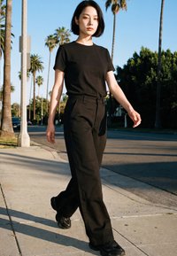 Young woman with short black hair walking on sunny sidewalk wearing a black t-shirt, black pants, and black sneakers with palm trees in background.