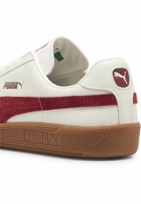 Białe skórzane sneakersy z czerwonym zamszowym paskiem, zaokrąglonym noskiem, gumową podeszwą, złotymi akcentami logo oraz wyściełanym kołnierzem.