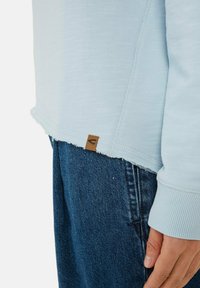 Jasnoniebieski sweatshirt o teksturowanym wykończeniu, z surowym dołem, ściągaczami rękawów i brązowym skórzanym akcentem na dole.