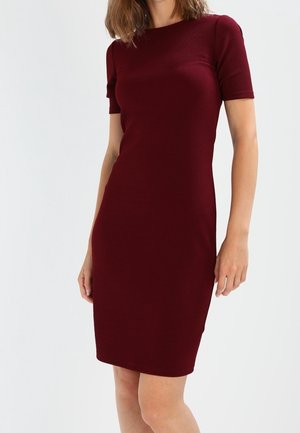 Femme portant une robe bordeaux ajustée, à manches courtes, longueur genou, debout devant un fond clair uni.