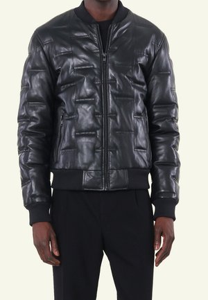 Mann trägt eine schwarze gesteppte Leder-Bomberjacke mit Reißverschlusstaschen sowie gerippten Bündchen und Saum, kombiniert mit schwarzen Hosen.