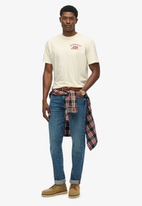 Camiseta gráfica beige con estampado de "Superdry", jeans de mezclilla azules, camisa de cuadros atada a la cintura y zapatos de cordones color marrón claro.