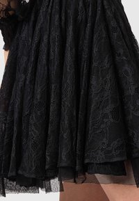 Robe en dentelle noire avec jupe plissée, présentant un motif floral en dentelle et un tulle superposé. Le tissu est transparent, créant une texture délicate.