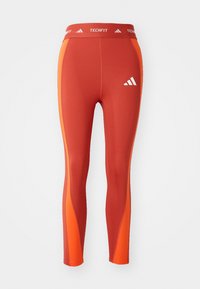 Punased sportlikud retuusid oranžide külgpaneelidega. Valmistatud venivast kangast, omavad laia vöökohta, millel on "TECHFIT" ja Adidas logo detailid.