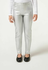 Silberne metallische Leggings mit glatter Textur und elastischem Bund, kombiniert mit schwarzen flachen Schuhen mit Schleifendetail.