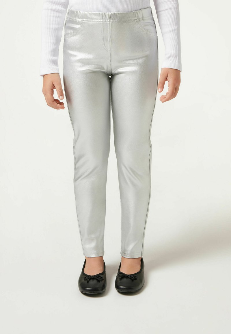 Silberne metallische Leggings mit glatter Textur und elastischem Bund, kombiniert mit schwarzen flachen Schuhen mit Schleifendetail.