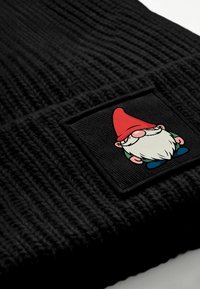 Schwarze gestrickte Mütze mit strukturiertem Muster. Verfügt über einen quadratischen Aufnäher mit einer bunten Gnom-Illustration, einschließlich rotem Hut und weißem Bart.