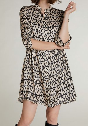 Donna che indossa un vestito beige e nero con motivo geometrico a bottoni, maniche lunghe arrotolate fino ai gomiti e stivali neri.