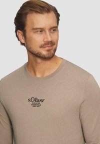 Beige langekm shirt met een ronde halslijn. Heeft een zwart geborduurd logo met de tekst "s.Oliver Elevated Essentials Since 1969." Glad textuur.