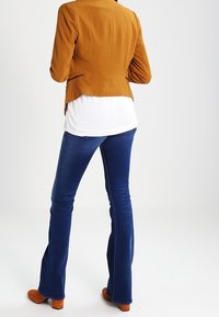 Giacca fitted gialla senape sopra una camicia bianca, abbinata a jeans a zampa d'elefante blu scuro e stivali con tacco marroni. Tessuto morbido e design strutturato.