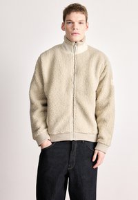 Bež flis jakna s visokim ovratnikom, patentnim zatvaračem sprijeda i rebrastim manšetama. Tekstura je mekana i pahuljasta, u kombinaciji s tamnim trapericama.