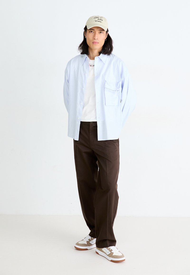 Hellblau gestreiftes Button-Down-Hemd über einem weißen T-Shirt, braune Hose und beigem Sneaker mit grauen Akzenten. Die Kappe hat einen Text.