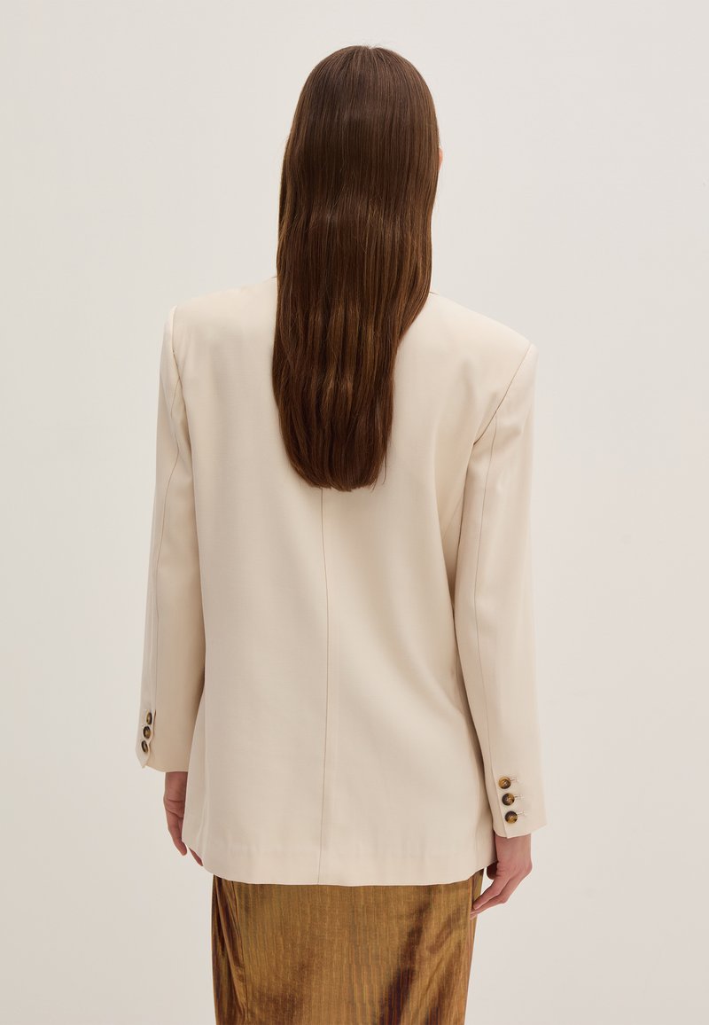 Blazer beige realizzato in tessuto liscio, con un taglio classico, maniche lunghe e tre bottoni dorati su ogni polsino. Vista posteriore mostrata.
