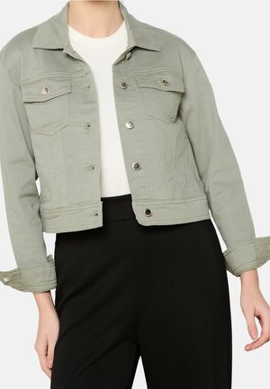 Frau trägt eine hellgrüne, kurze Jeansjacke über einem weißen Oberteil und schwarzen Hosen, wobei die Jackenärmel teilweise hochgekrempelt sind.