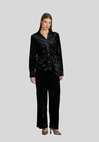 Zwarte fluwelen blouse met knoopsluiting en gathered detail, gecombineerd met bijpassende wijde broek. Model draagt zilveren hakken met bandjes.