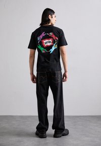 Black cotton t-shirt featuring a colorful graphic design on the back with playful patterns and text. Paired with black, wide-leg jeans.