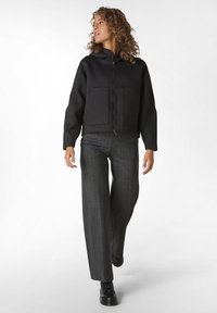 Felpa nera con zip e due tasche frontali, abbinata a pantaloni larghi grigi a righe e scarpe nere, con una texture liscia.