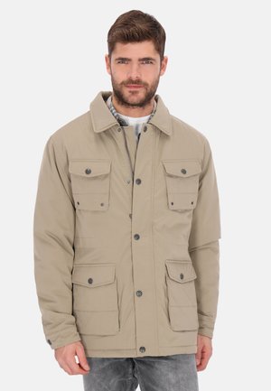 Ung mand iført en beige knappet jakke med fire forlommer, stående mod en ensfarvet lys baggrund.