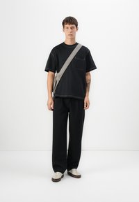 Svart t-skjorte med kort erm og lomme på brystet, med kontrastfargede sømmer, sammen med løse svarte bukser og lysfargede joggesko.