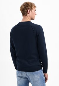 Middendingsblauwe gebreide trui met geribde halslijn en boorden, met een eenvoudig ontwerp en een iets gestructureerd oppervlak, gedragen met lichtblauwe jeans.
