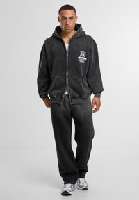 Dunkelgrauer Zip-Hoodie mit weicher Textur, der ein Logo auf der Brust und große Fronttaschen hat. Kombiniert mit passenden Jogginghosen.