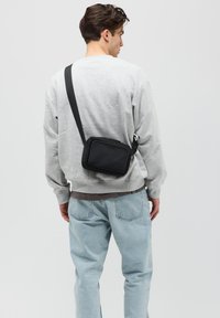 Schwarze Stoff-Crossbody-Tasche mit einer vorderen Reißverschlusstasche, angenhängtem Schultergurt und glatter Textur, getragen von einer Person in einem grauen Sweatshirt.