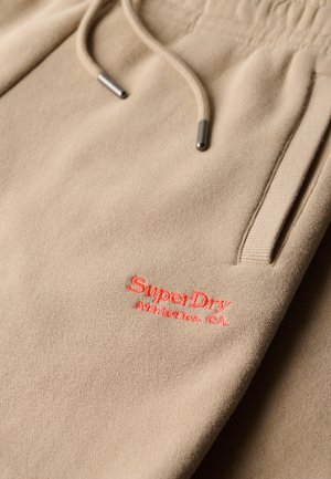 Pantaloni della tuta beige con vita elastica, coulisse e tasche anteriori. Presentano il logo ricamato "SuperDry Athletics Co." in rosso.