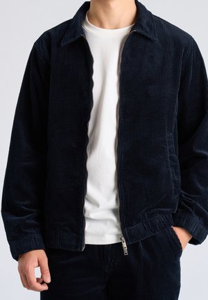 Mann trägt dunkelblaue Cordjacke über weißem T-Shirt, mit einer Hand in der Tasche und der anderen locker seitlich.